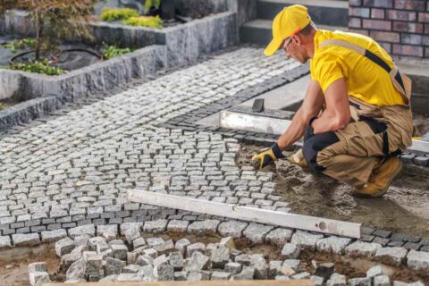 A construction worker in a yellow shirt and cap is meticulously placing cobblestones to create a pathway in a landscaped garden.